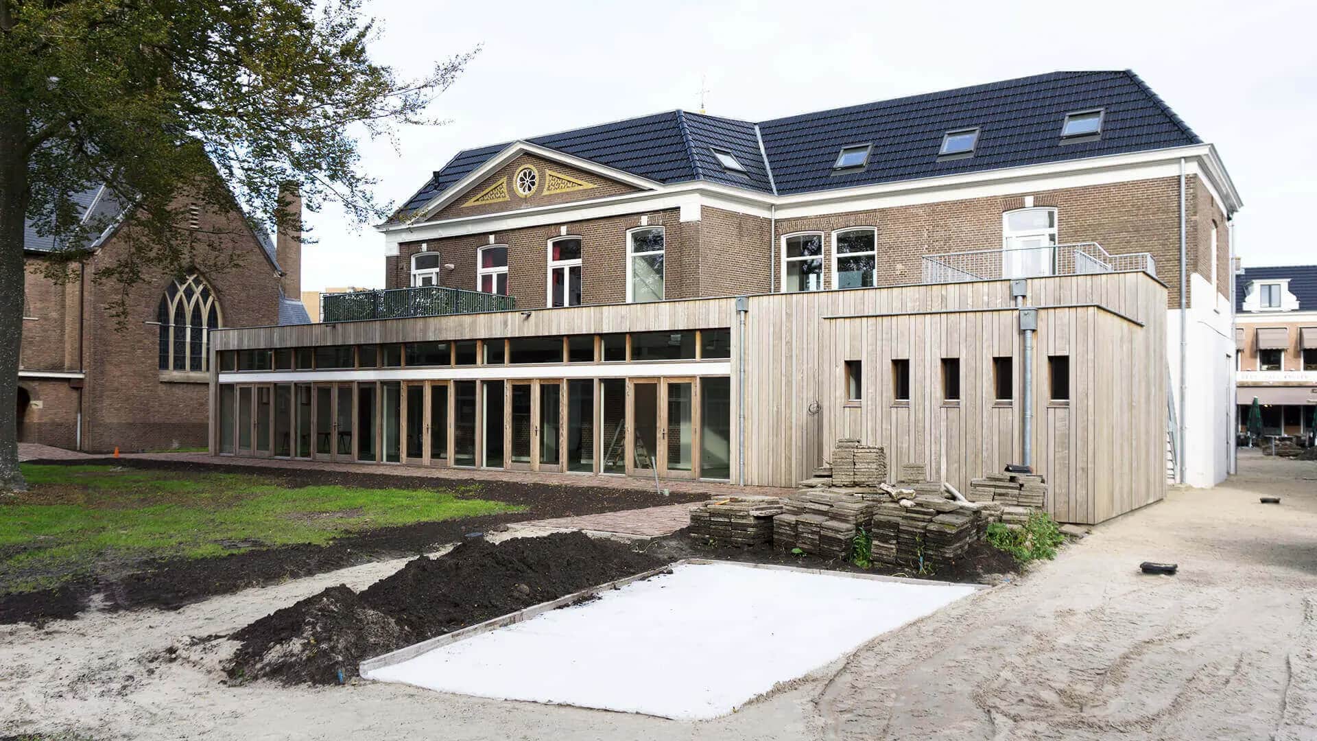 Ontmoetingscentrum Heerenveen: modern uitbreiding met houten gevel naast historische bakstenen kerk, onder constructie